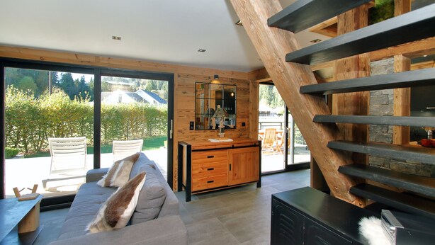 A modern living room with a large window, a wooden staircase, and a built-in kitchen area.