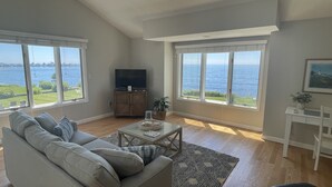 Een woonkamer met een bank, een salontafel en een televisie. Grote ramen bieden uitzicht op zee.