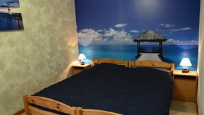 Een slaapkamer met een bed, twee nachtkastjes met lampen, een bijzettafeltje en een muurschildering van een strand met een pier en een rieten gebouw.