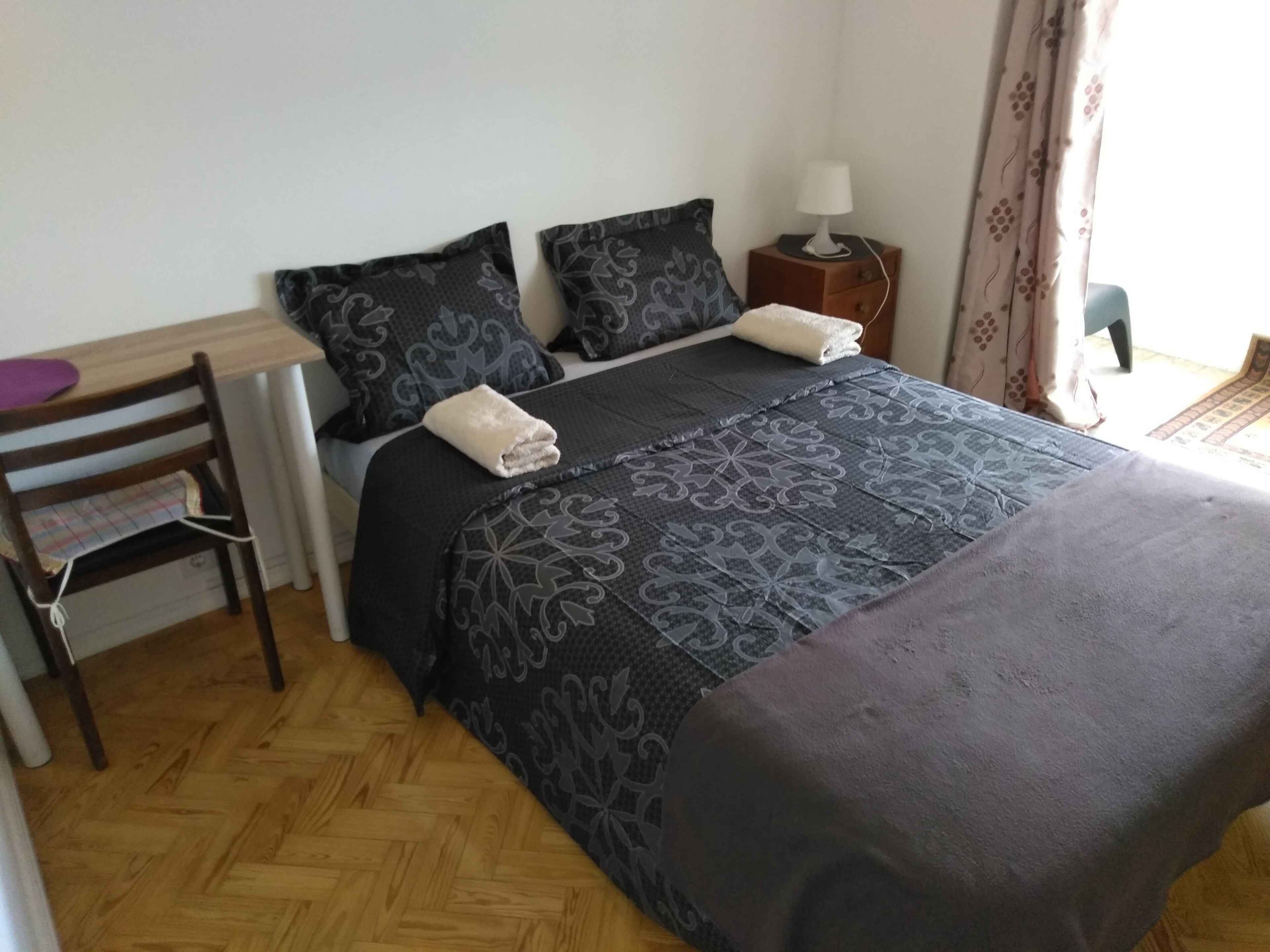 A neatly made bed with a patterned bedspread, a bedside table with a lamp, and a chair with a cushion.