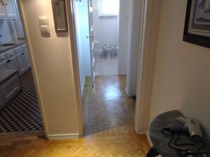 A narrow hallway with a herringbone floor pattern leading to a bathroom with a toilet and bidet. A hairdryer is visible on the floor to the right. - Voyage 63 (Lisbon)