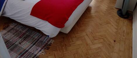 A neatly made bed with a blue and red blanket, a wooden floor, and a standing heater.