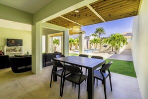 A modern outdoor dining area with a wooden pergola, a pool, and palm trees.