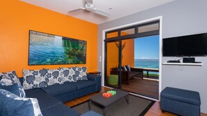 A modern living room with a sofa, coffee table, and a TV. A large window offers a view of the ocean. - Panama Beachfront Apartments Rarotonga (Rarotonga)