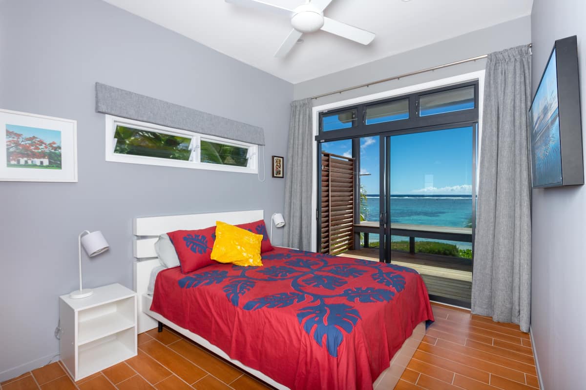 A bedroom with a bed, a nightstand, a ceiling fan, a window with a view of the sea, and a picture on the wall.