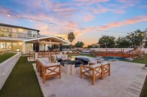 Un patio al aire libre con asientos y un hogar para fuego, rodeado por una piscina y un jardín ajardinado.
