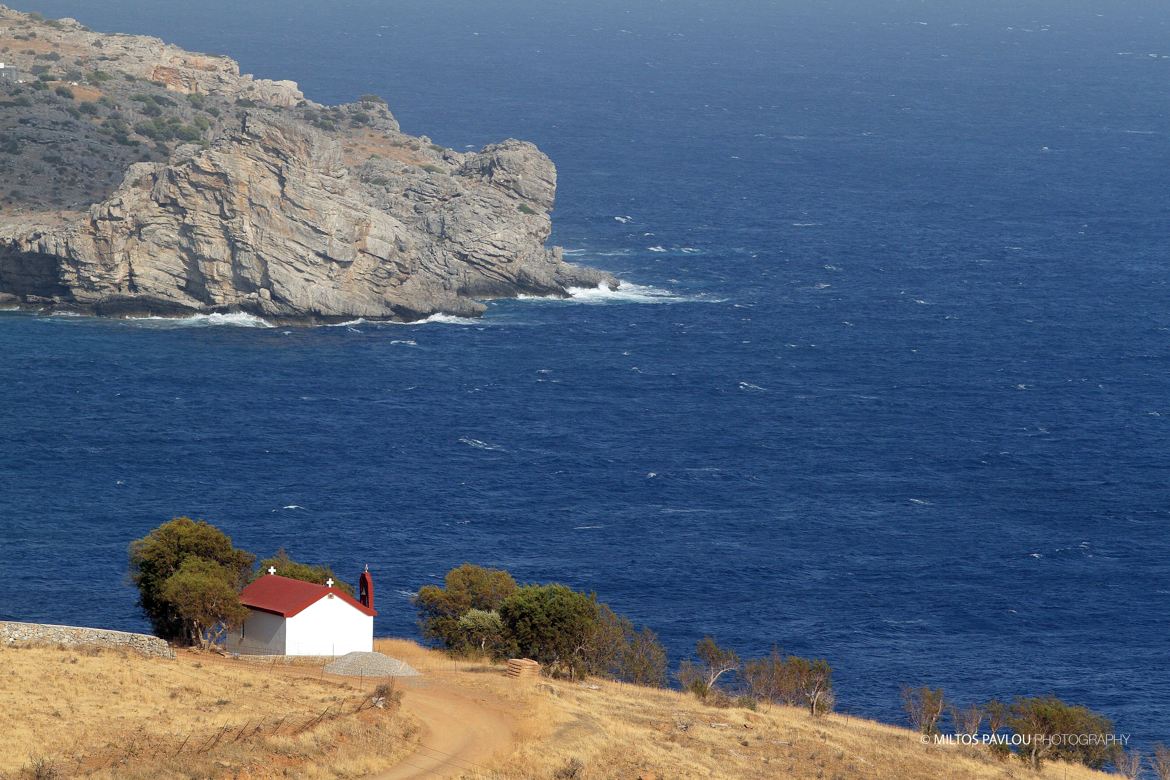 A coastal scene with a small white building with a red roof, situated on a hillside with a rocky cliff in the background and the sea in the foreground.