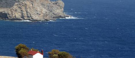 A coastal scene with a small white building with a red roof, situated on a hillside with a rocky cliff in the background and the sea in the foreground.