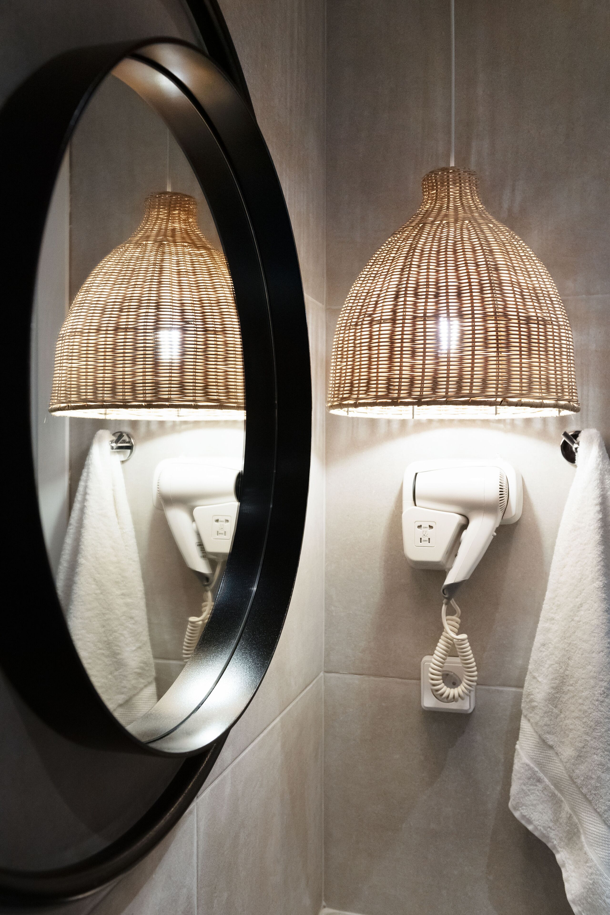A bathroom with a round mirror, a wall-mounted hairdryer, and a woven lamp.