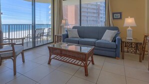Un salon avec un canapé bleu, une table basse en verre et un banc en osier. Deux lampes sont posées sur des tables d’appoint, et de grandes fenêtres offrent une vue sur la plage.