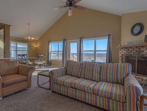 A living room with a striped sofa, a brown armchair, and a dining area with a table and chairs. There is a fireplace with a 'BEACH' sign above it and a clock on the wall.
