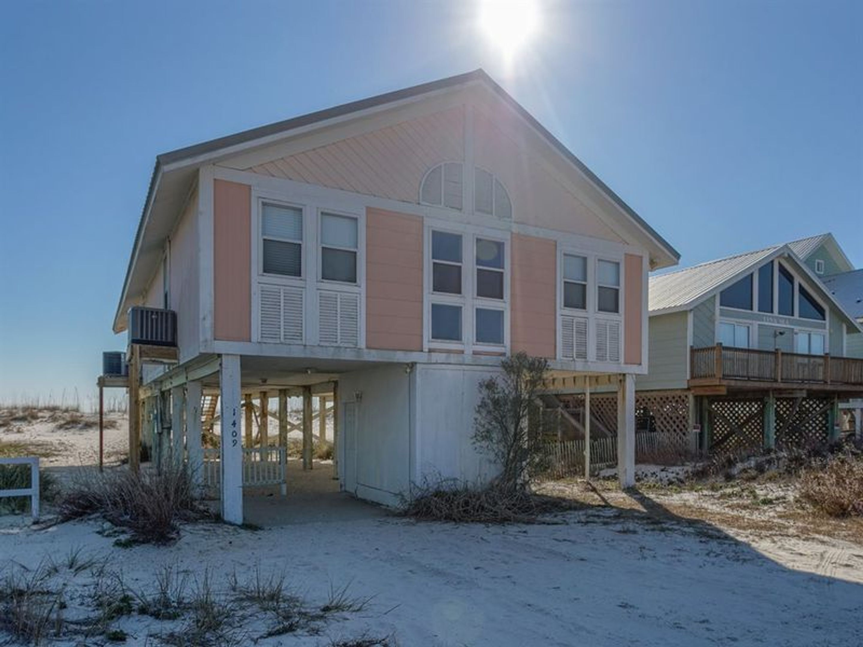 Une maison de plage à deux étages, dotée d’un porche couvert, de grandes fenêtres et située sur une plage de sable.