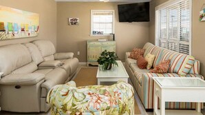 A living room with a beige sofa, a striped couch, a floral ottoman, a coffee table, a ceiling fan, a TV mounted on the wall, and a window with blinds. - Red Robyn by Meyer Vacation Rentals (Gulf Shores)