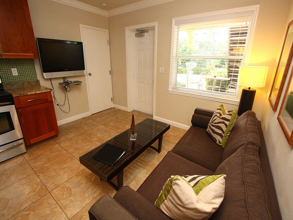 A living room with a brown sofa, a glass coffee table, a flat-screen TV mounted on the wall, and a window with blinds.