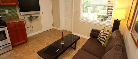 A living room with a brown sofa, a glass coffee table, a flat-screen TV mounted on the wall, and a window with blinds.