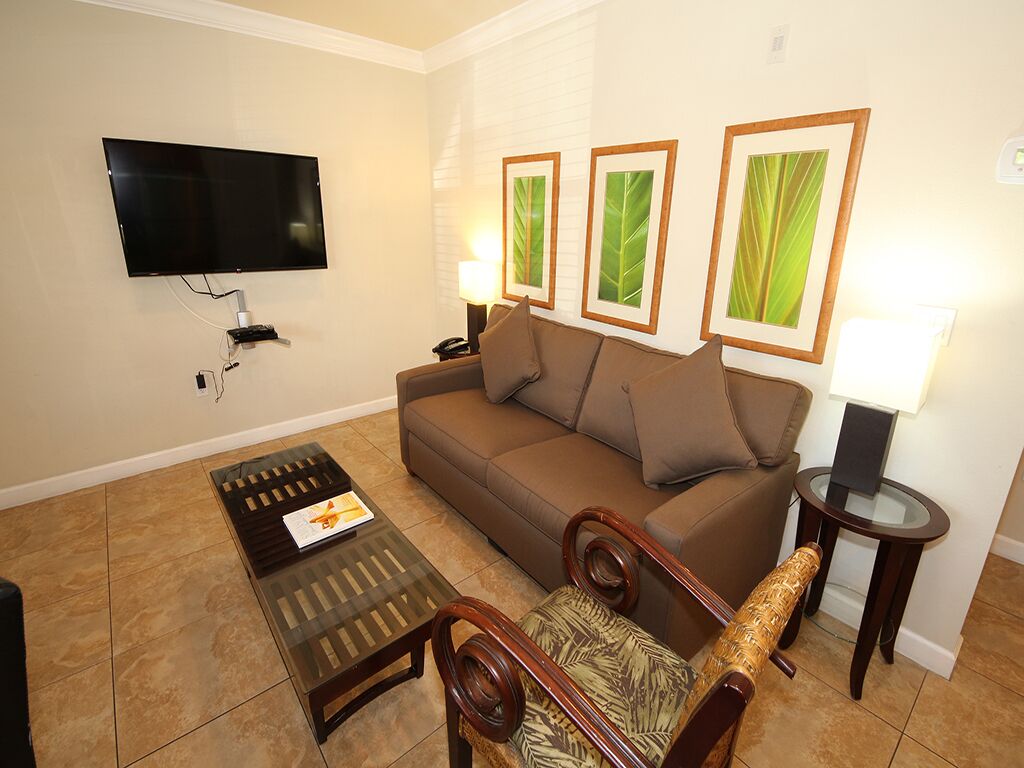 A living room with a flat-screen TV mounted on the wall, a brown sofa, a glass coffee table, and a patterned armchair.