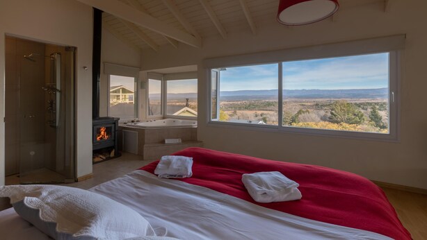 A bedroom with a large bed, a red blanket, a fireplace, and a view of the countryside. - La Fleur Loft Boutique Spa (Villa General Belgrano)