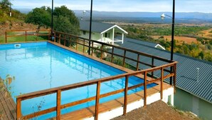 A rectangular pool with a wooden deck and metal railings, surrounded by a green landscape and a house with a red roof. - La Fleur Loft Boutique Spa (Villa General Belgrano)