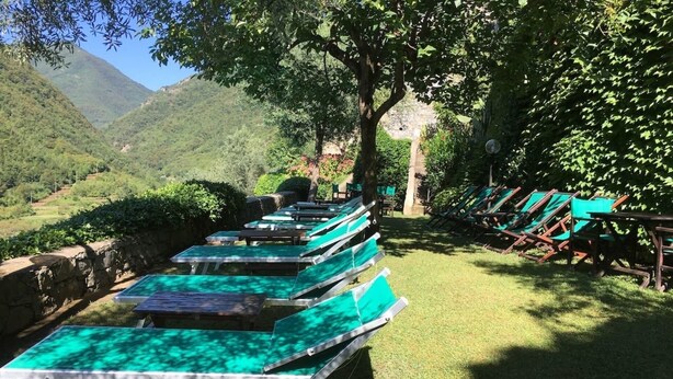 A row of green lounge chairs and tables arranged outdoors, with a stone wall and trees in the background.