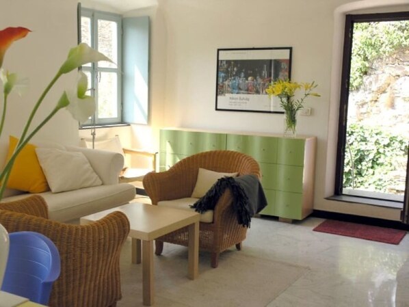Una sala de estar con sofá, sillas, una mesa y un aparador verde. Se ve un ventanal con vistas a la vegetación.