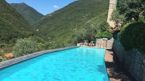 Una piscina rectangular con agua azul cristalina, rodeada por una terraza de madera, con montañas verdes y frondosas al fondo.