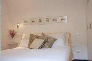 A neatly made bed with white bedding and patterned pillows, a small bedside table with a vase of flowers, and a wall with framed pictures. - 2 Bed House Sleeps 5 - Terrace - Communal Pool (Castelbianco)
