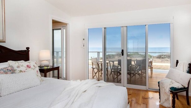 A bedroom with a large bed, a nightstand, and a view of the beach through a sliding glass door. - PORT WILLUNGA DREAMING a beach front experience (Port Willunga)