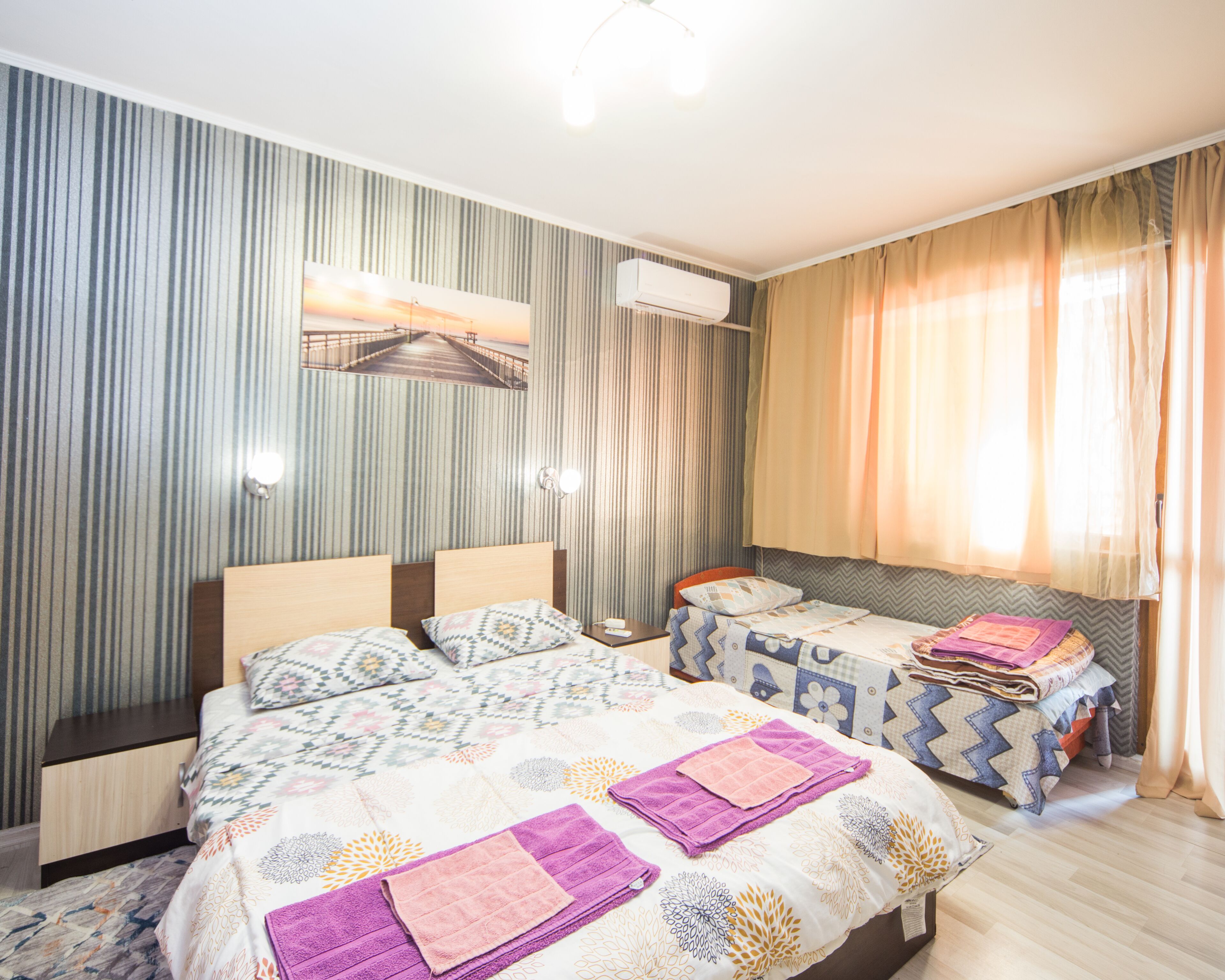 A hotel room with two beds, striped wallpaper, an air conditioning unit, a painting of a sunset, and a window with curtains.
