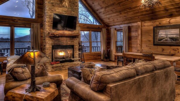L’intérieur chaleureux d’un chalet en bois, avec une cheminée en pierre, des meubles en cuir et de grandes fenêtres offrant une vue imprenable sur le paysage.