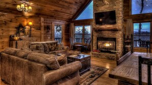 L’intérieur chaleureux d’un chalet en bois comprend un grand canapé en cuir, une cheminée en pierre et un téléviseur fixé au mur.