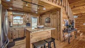 L’intérieur d’un chalet rustique, avec des murs et des planchers en bois, un coin cuisine doté de comptoirs en granit, une grande fenêtre et un coin repas avec des tabourets.
