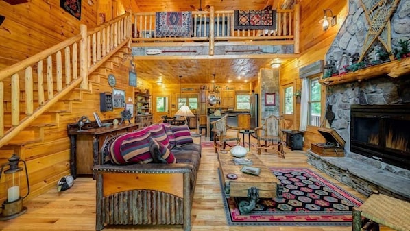 A log cabin interior with a wooden staircase, a fireplace, a dining area, and a kitchen.