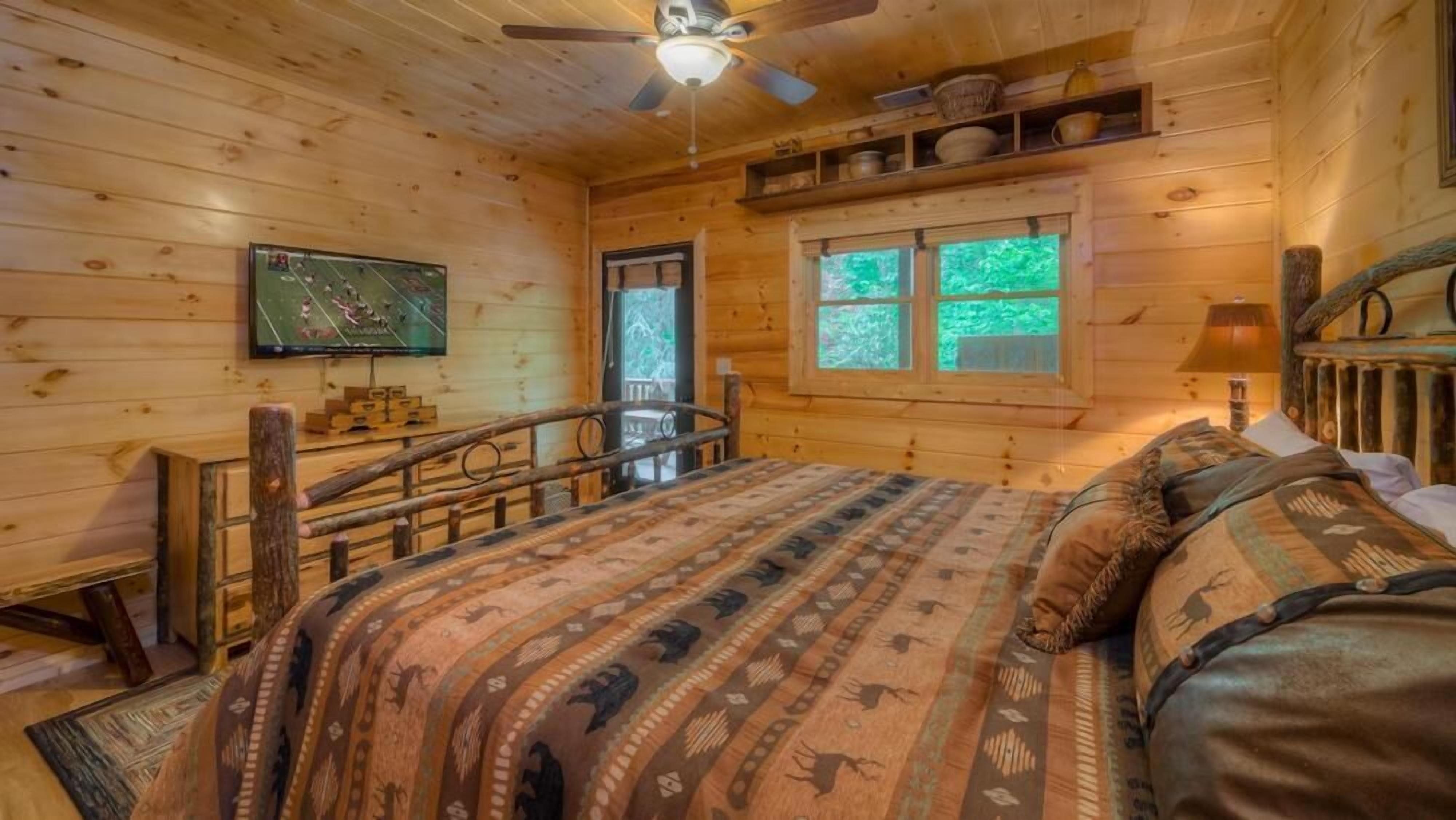 A wooden cabin bedroom with a bed, a ceiling fan, a television, and a window with a view of greenery.