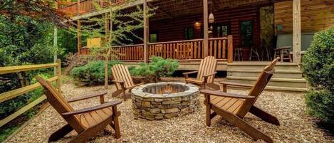 A cabin with a stone fire pit surrounded by Adirondack chairs, a wooden deck, and a stone wall.