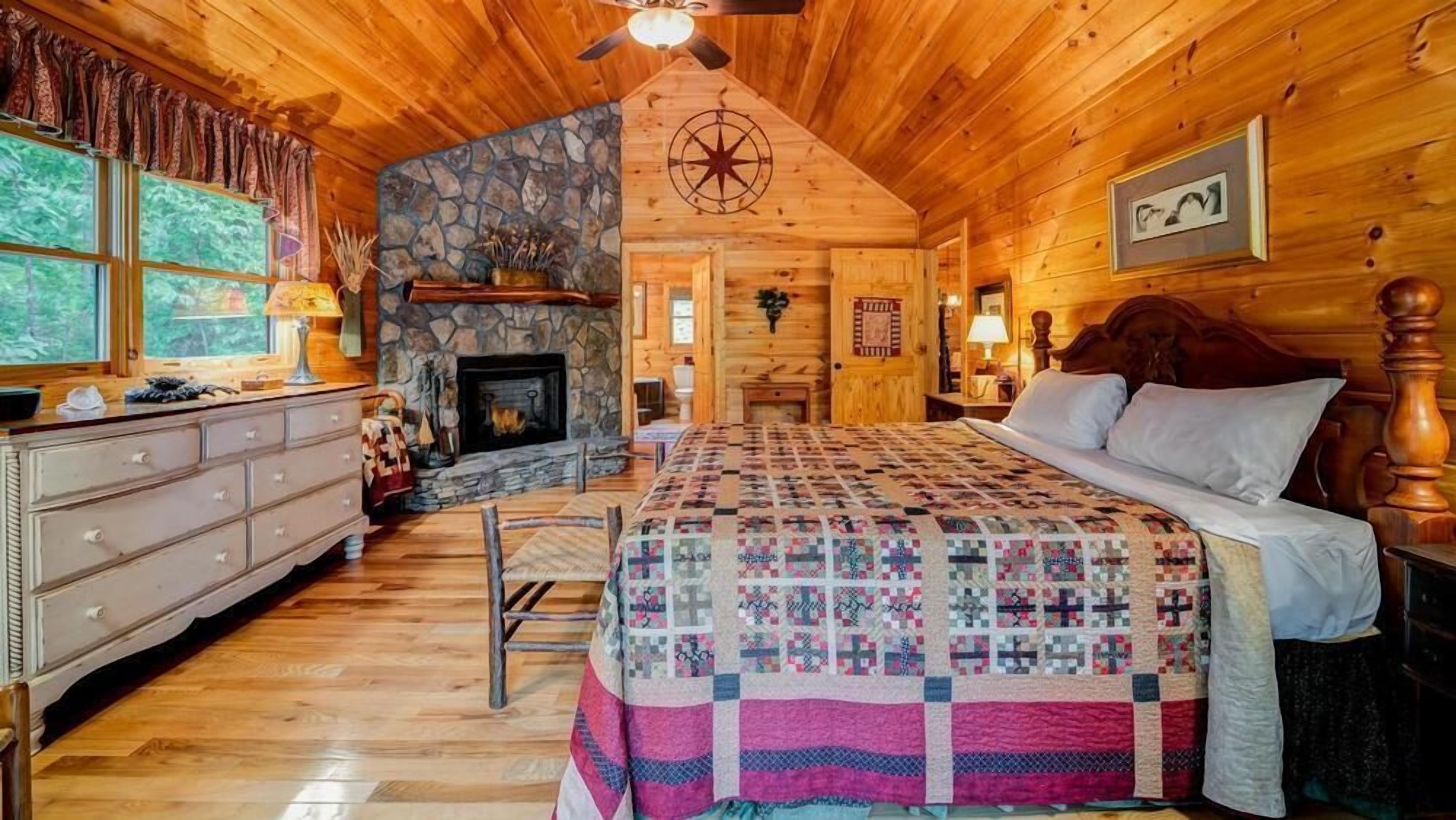 A cozy log cabin bedroom with a stone fireplace, a quilted bedspread, and a wooden dresser.