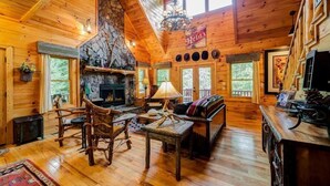 L’intérieur chaleureux d’un chalet avec une cheminée en pierre, des meubles en bois et de grandes fenêtres offrant une vue sur le paysage extérieur.