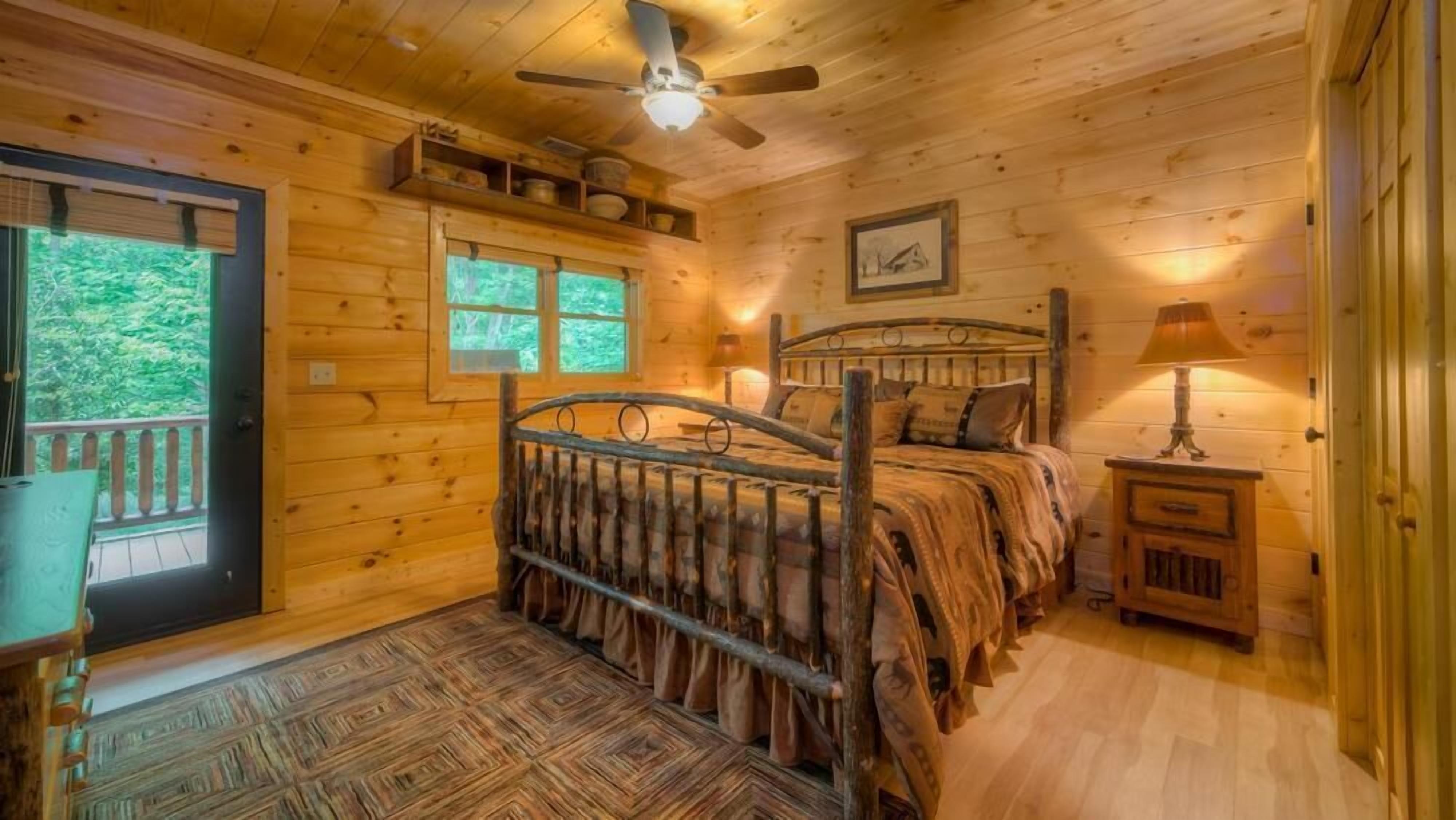 A cozy wooden cabin bedroom with a bed, a ceiling fan, a window, and a door.