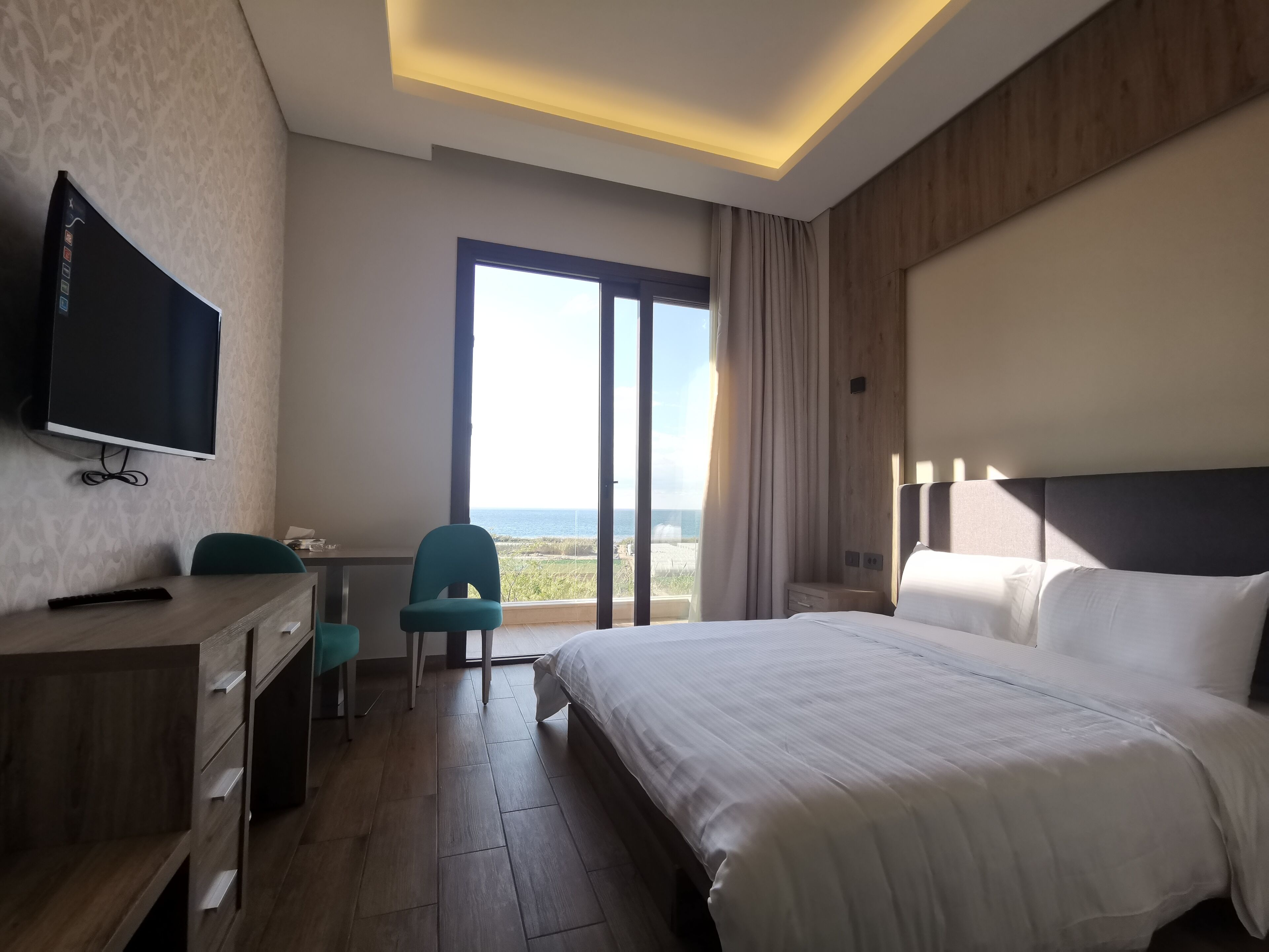 Habitación de hotel con una cama grande, un escritorio con una silla, televisión y vista al mar a través de un ventanal amplio.