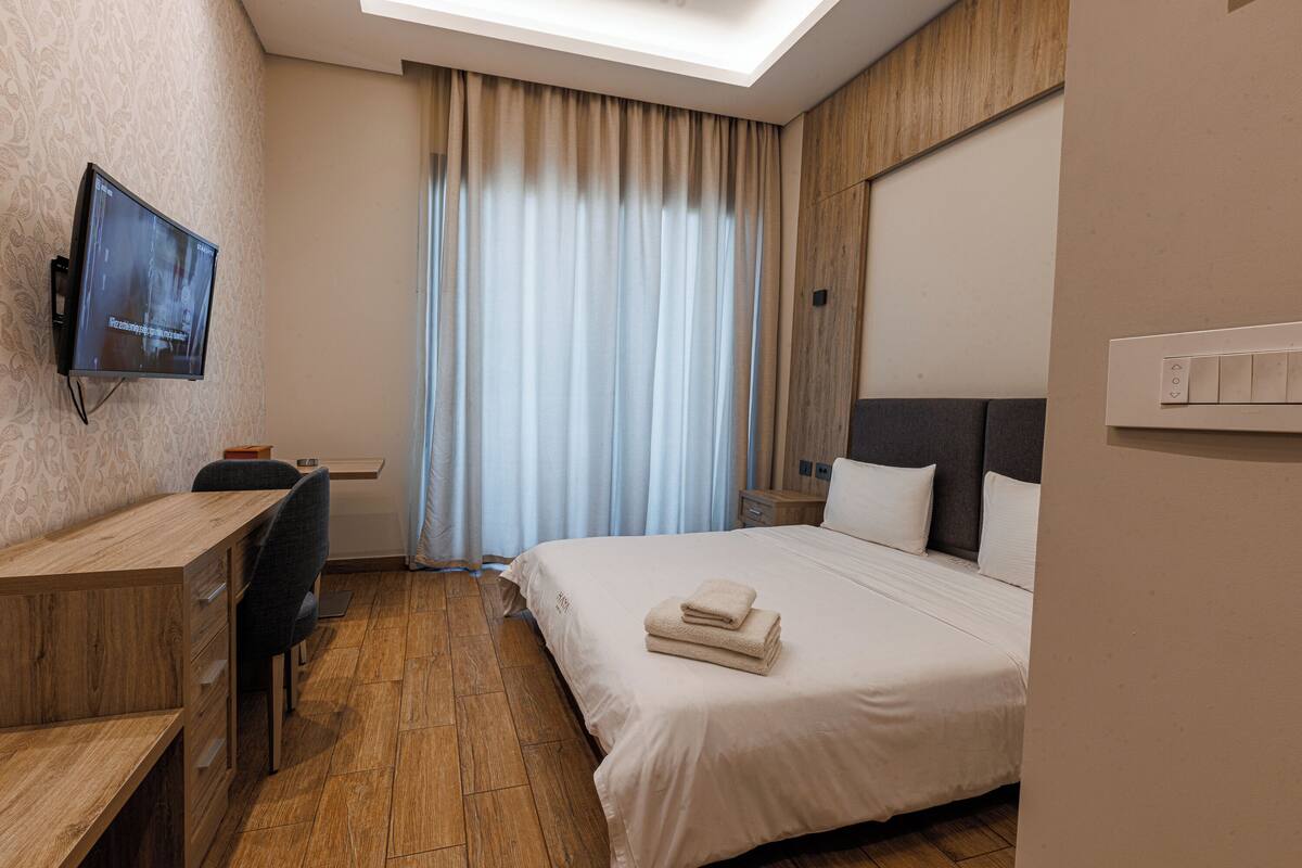 A hotel room with a wooden desk, a bed with white linens, a flat-screen TV mounted on the wall, and a large window with curtains.