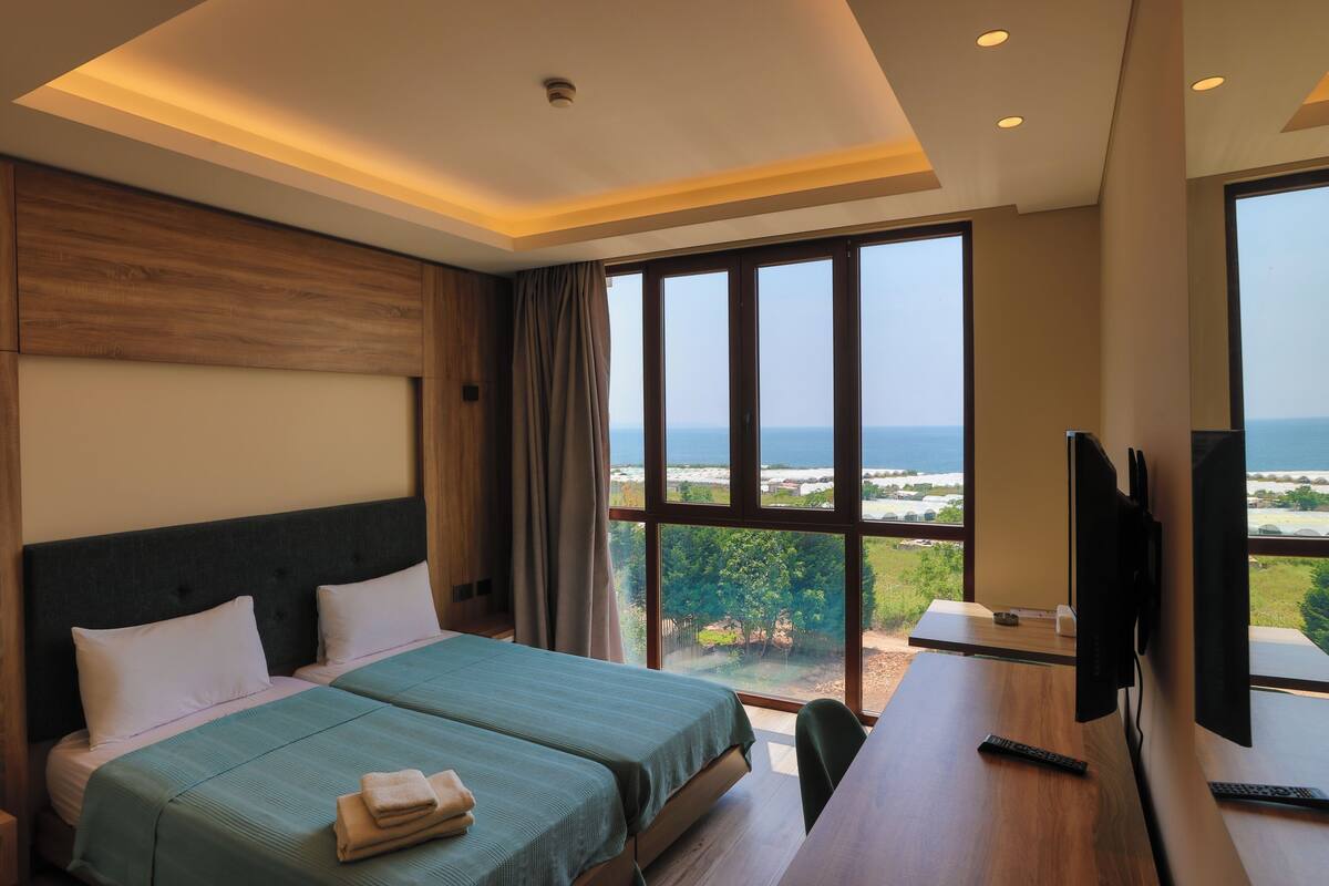 A hotel room with a bed, a desk, a TV, and a large window offering a view of the sea.