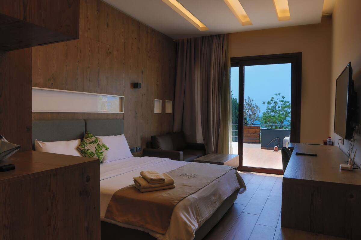 A modern hotel room with a large bed, wooden furniture, a desk, and a balcony with a view.