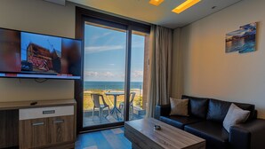 A modern living room with a large TV, a wooden coffee table, a black leather sofa, and a view of the ocean through large glass doors. - Haya hotel & spa (Tabarja)