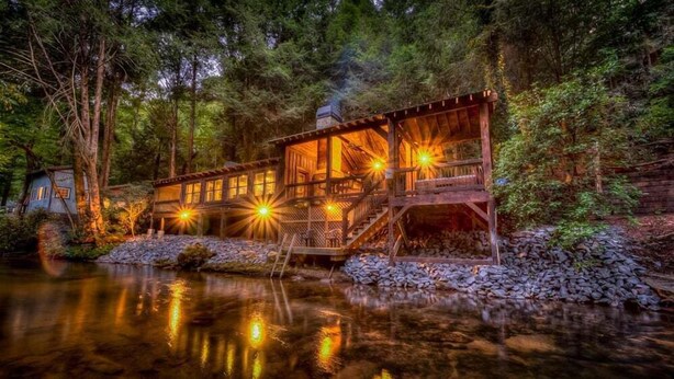 Una cabaña rústica con una terraza de madera y base de piedra, rodeada de un bosque denso y un lago tranquilo.