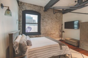 A bedroom with a brick wall, a window, a bed with pillows, a bedside lamp, a television, and a wardrobe.