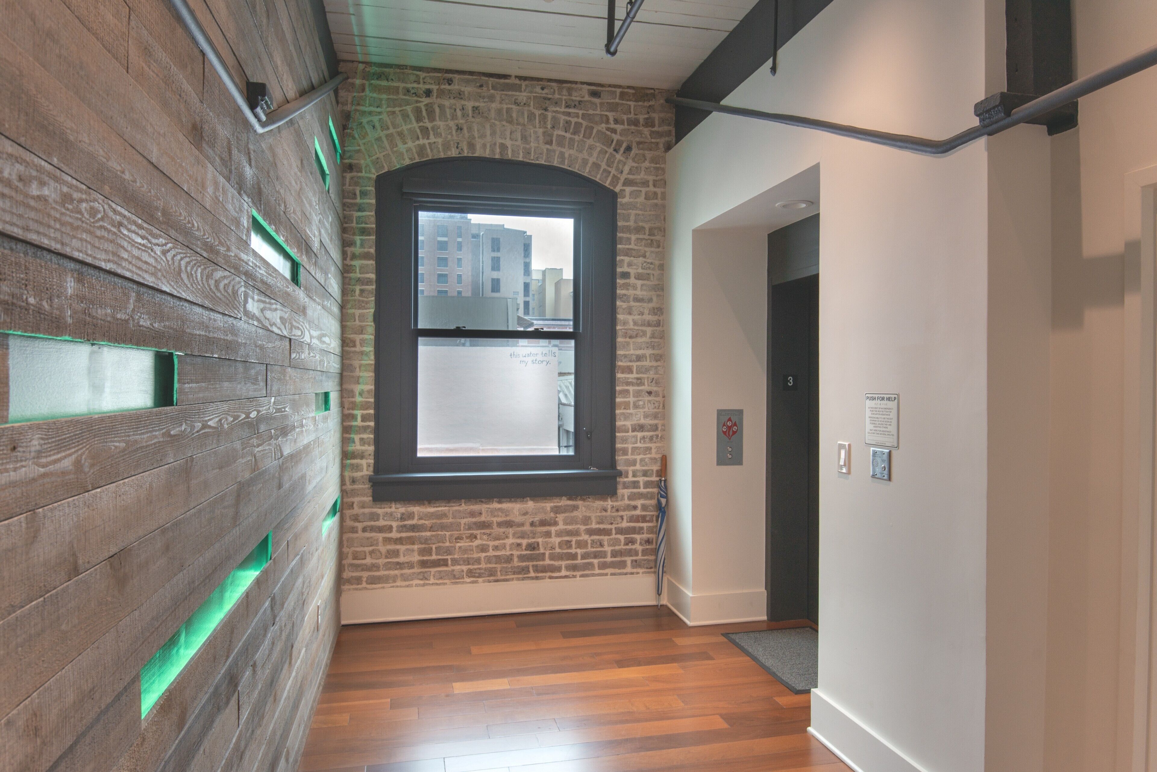 A hallway with a brick wall, wooden floor, and a window with a view of buildings outside.