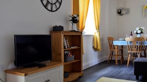 A living room with a flat-screen TV, wooden bookshelf, dining table with chairs, and a clock on the wall. - Woodlands View (Halesworth)