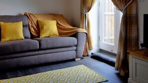 A living room with a grey sofa, yellow cushions, a yellow patterned rug, plaid curtains, and a pendant light. - Woodlands View (Halesworth)