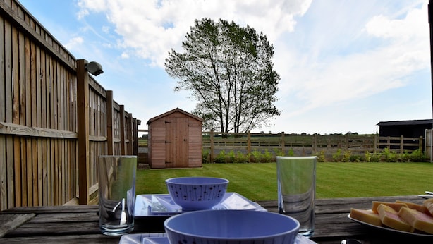 A wooden deck with a picnic setup including a plate of food, two bowls, and glasses. - Woodlands View (Halesworth)