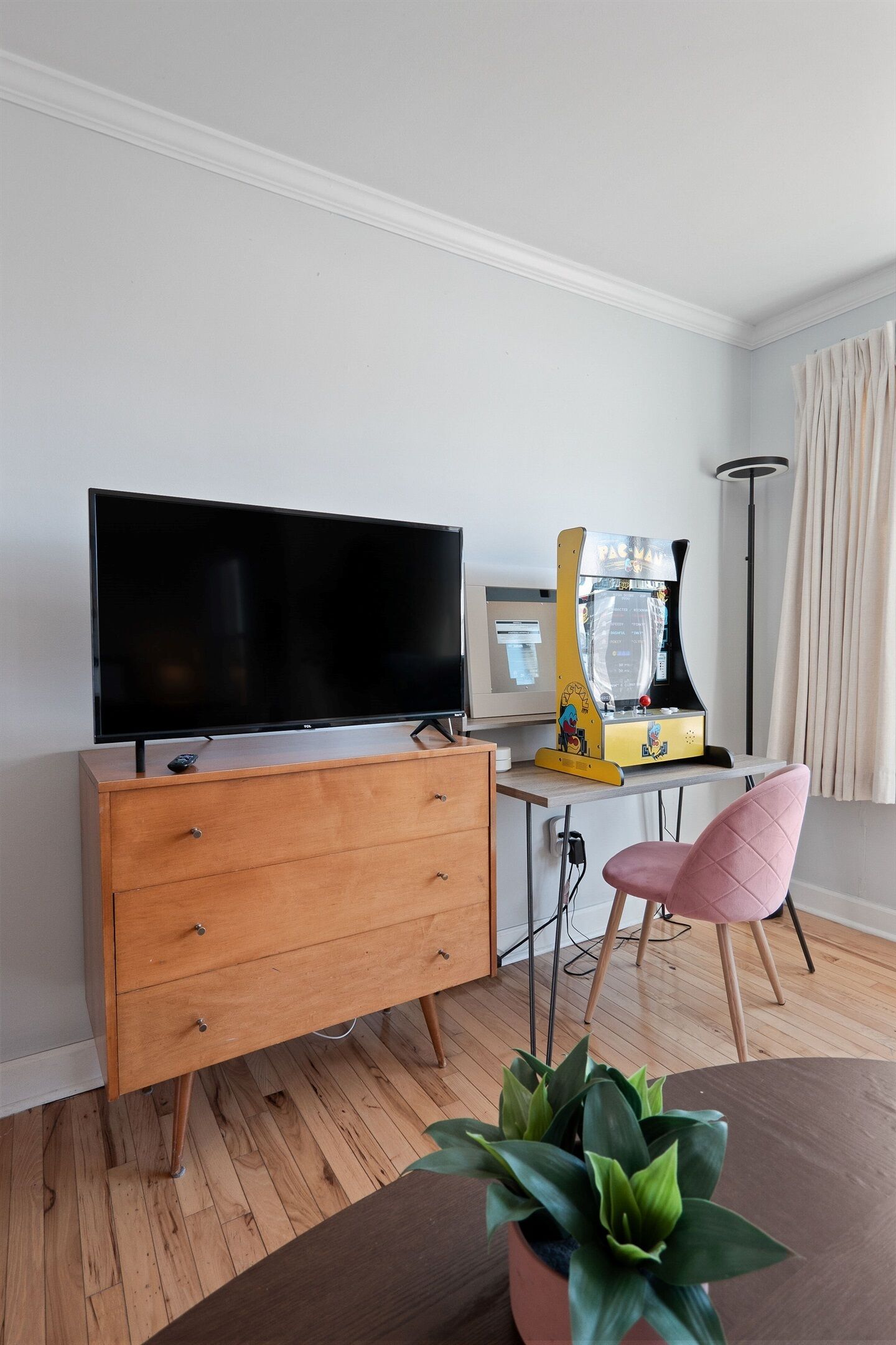 A room with a TV, wooden dresser, desk with arcade game, pink chair, and a potted plant.