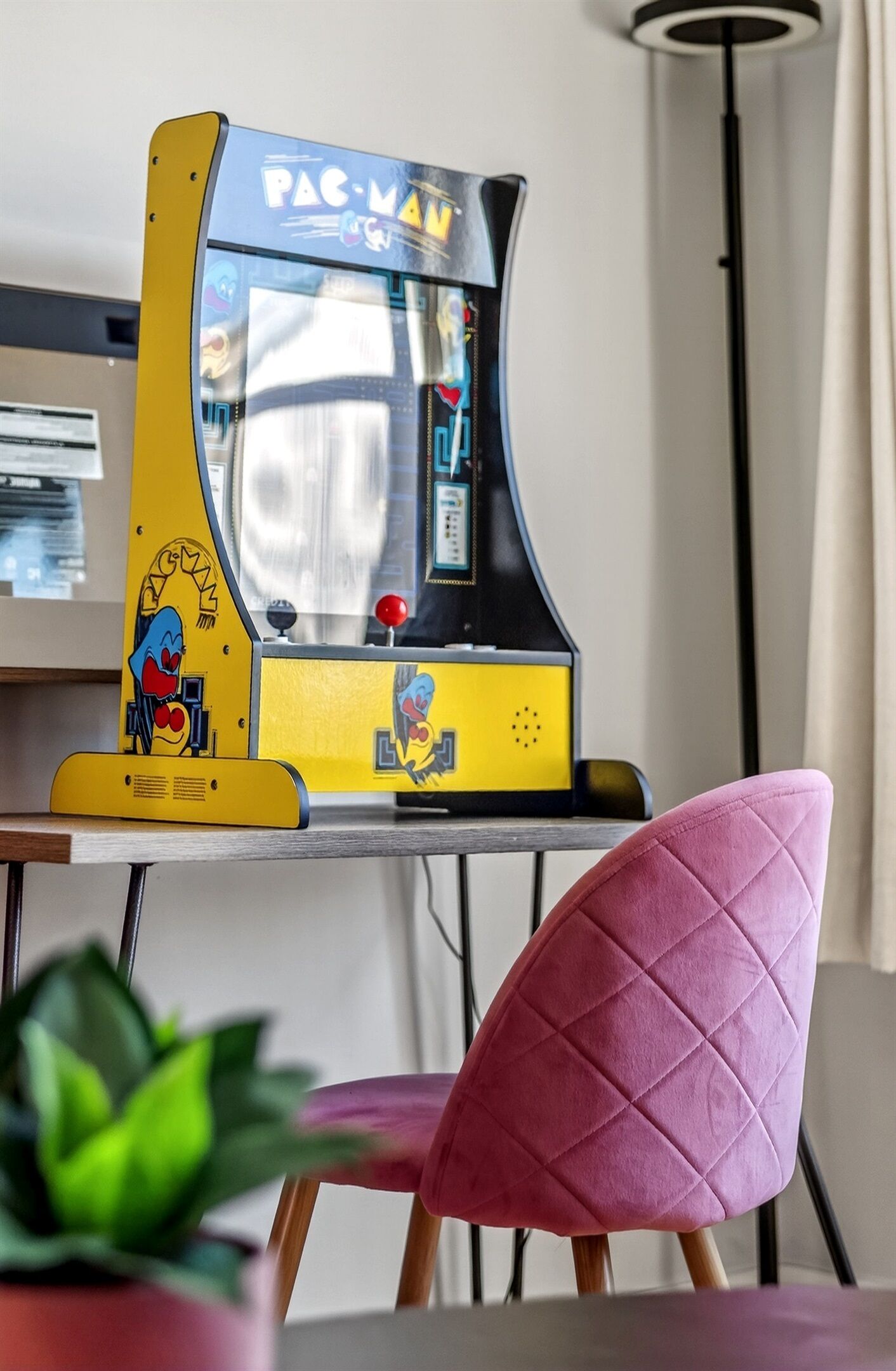 A Pac-Man arcade machine on a table with a pink chair in the foreground.